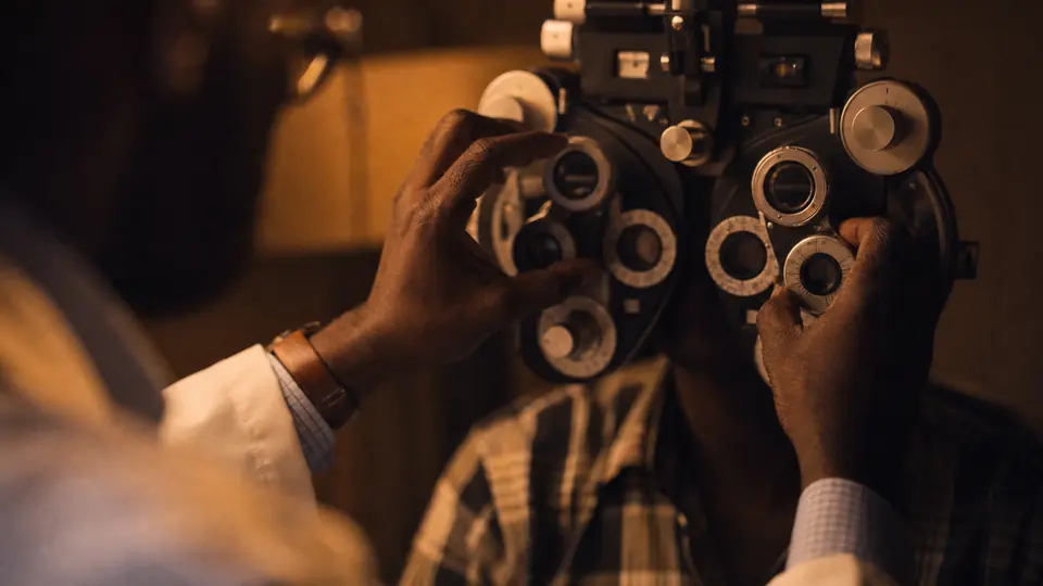An optometrist's hands at a phoropter in a dim exam room, lit by one warm lamp